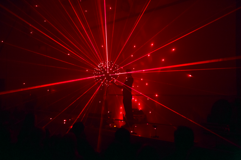 En diskokula som det skuts rött ljus på vilket får effekten av att hela rummeet är fullt av röda ljusstrålar. En man står också vid den stora kulan och vänder på den. 