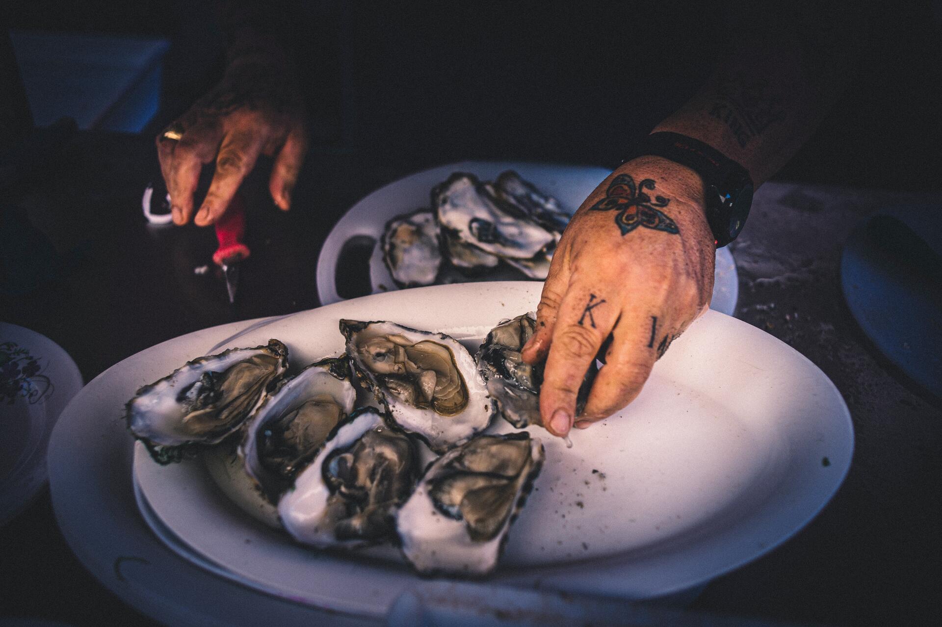 Tattooed hands opening a tray of oysters.